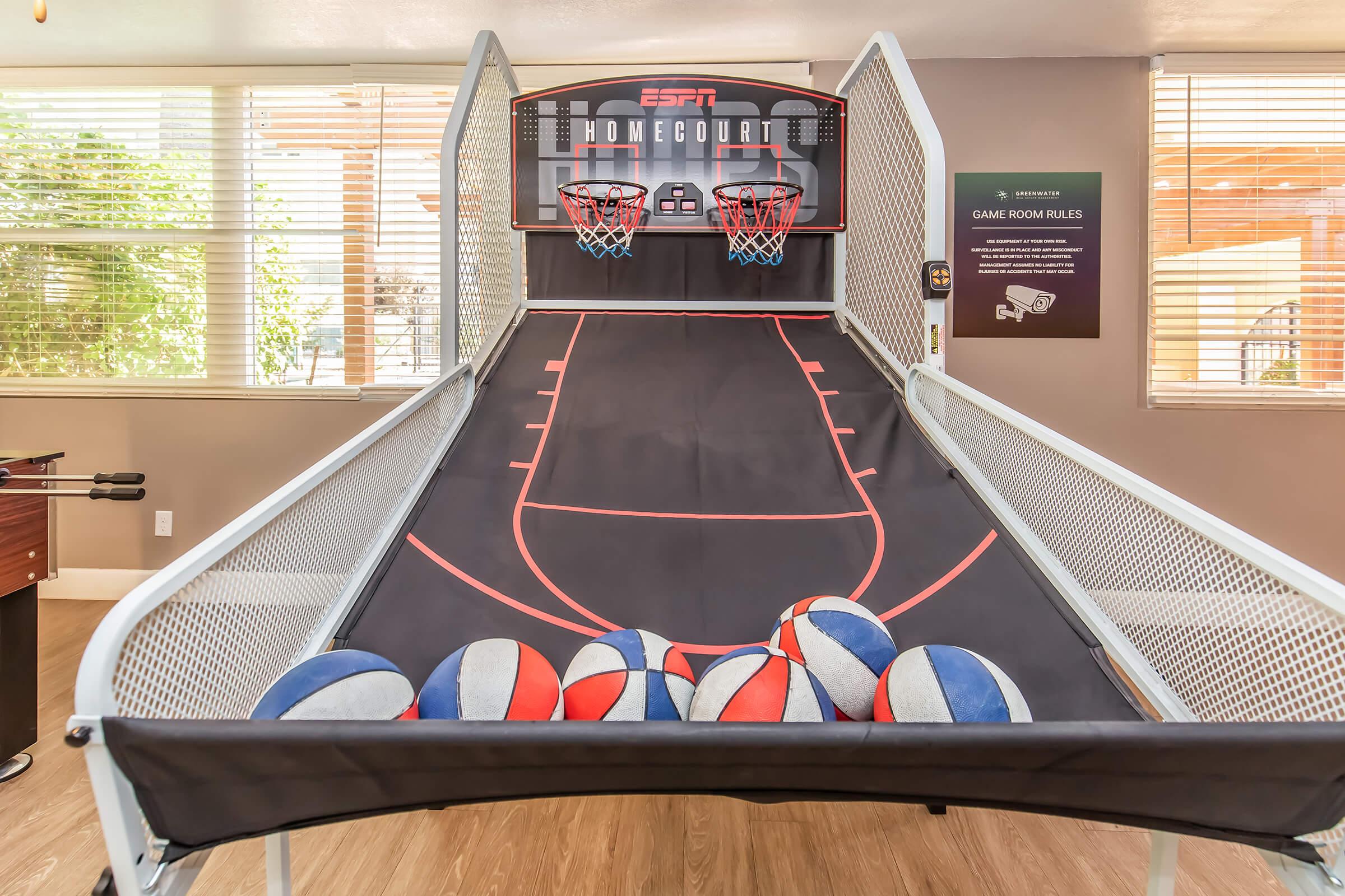 A basketball arcade game with two hoops and a black playing surface is set up in a well-lit room. Several colorful basketballs are scattered at the base of the game. Windows allow natural light to fill the space, enhancing the inviting atmosphere. A sign with game rules is visible on the wall.