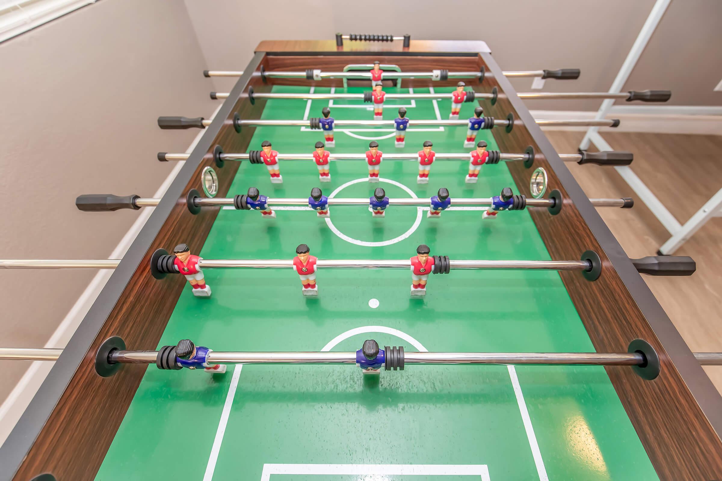 A top-down view of a foosball table featuring a green playing field. The table has red and blue player figures positioned on rods, with a scoring system at each end. The wooden frame and the arrangement of players indicate an active game setup.