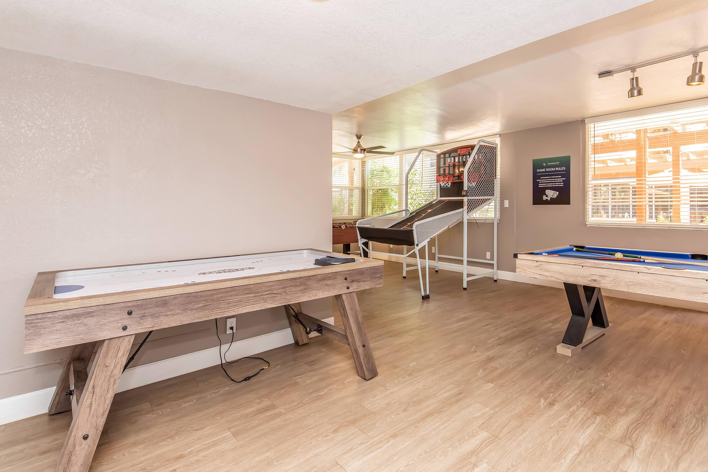 An indoor recreation room featuring a pool table, air hockey table, and a dartboard area. Large windows provide natural light, and modern decor enhances the inviting atmosphere. The flooring is light-colored, complementing the neutral wall tones.