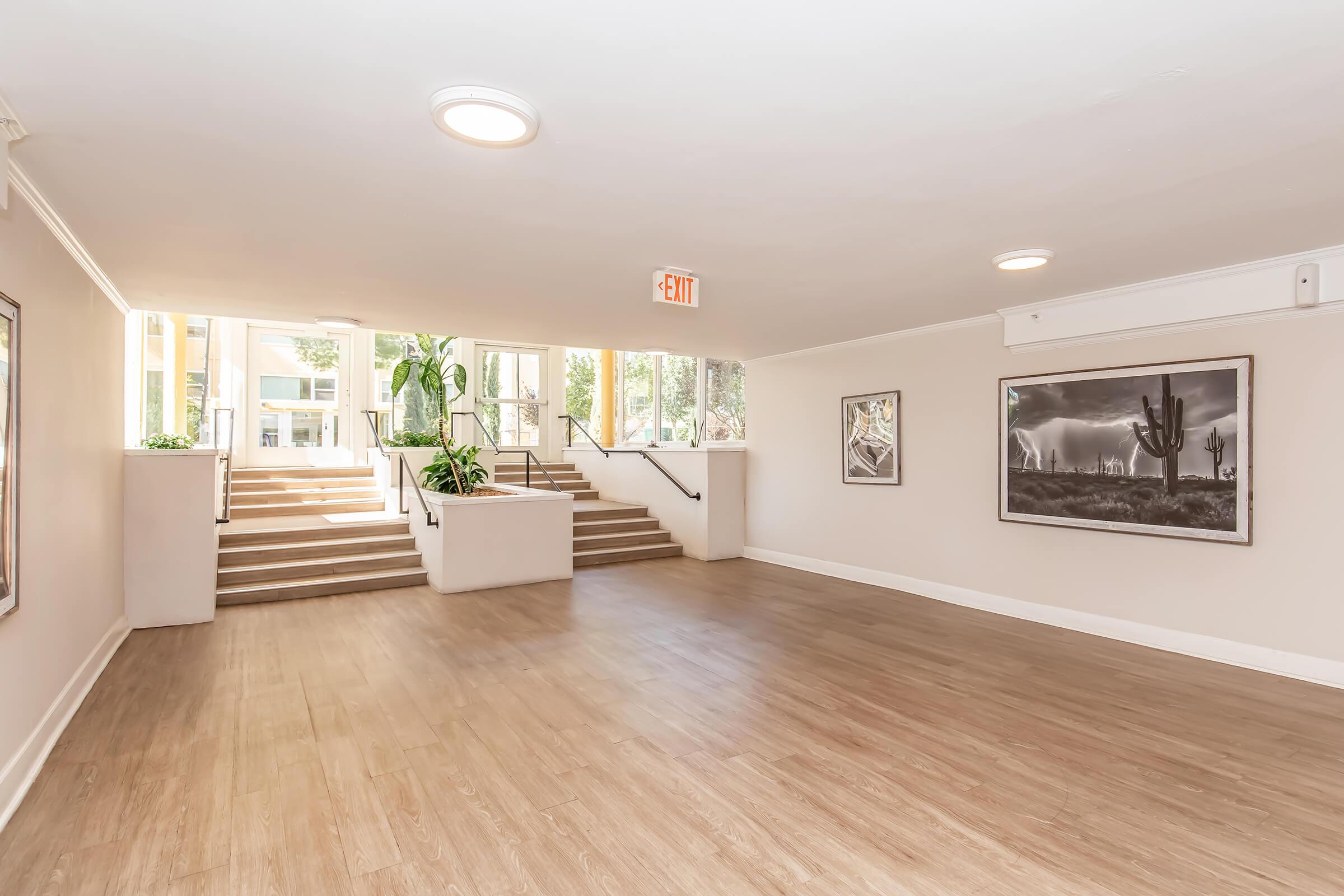 Interior view of a spacious hallway with wooden flooring, featuring framed black-and-white photographs on the walls. Natural light comes through large windows at the far end, leading to a staircase with potted plants on either side. The overall atmosphere is modern and inviting.