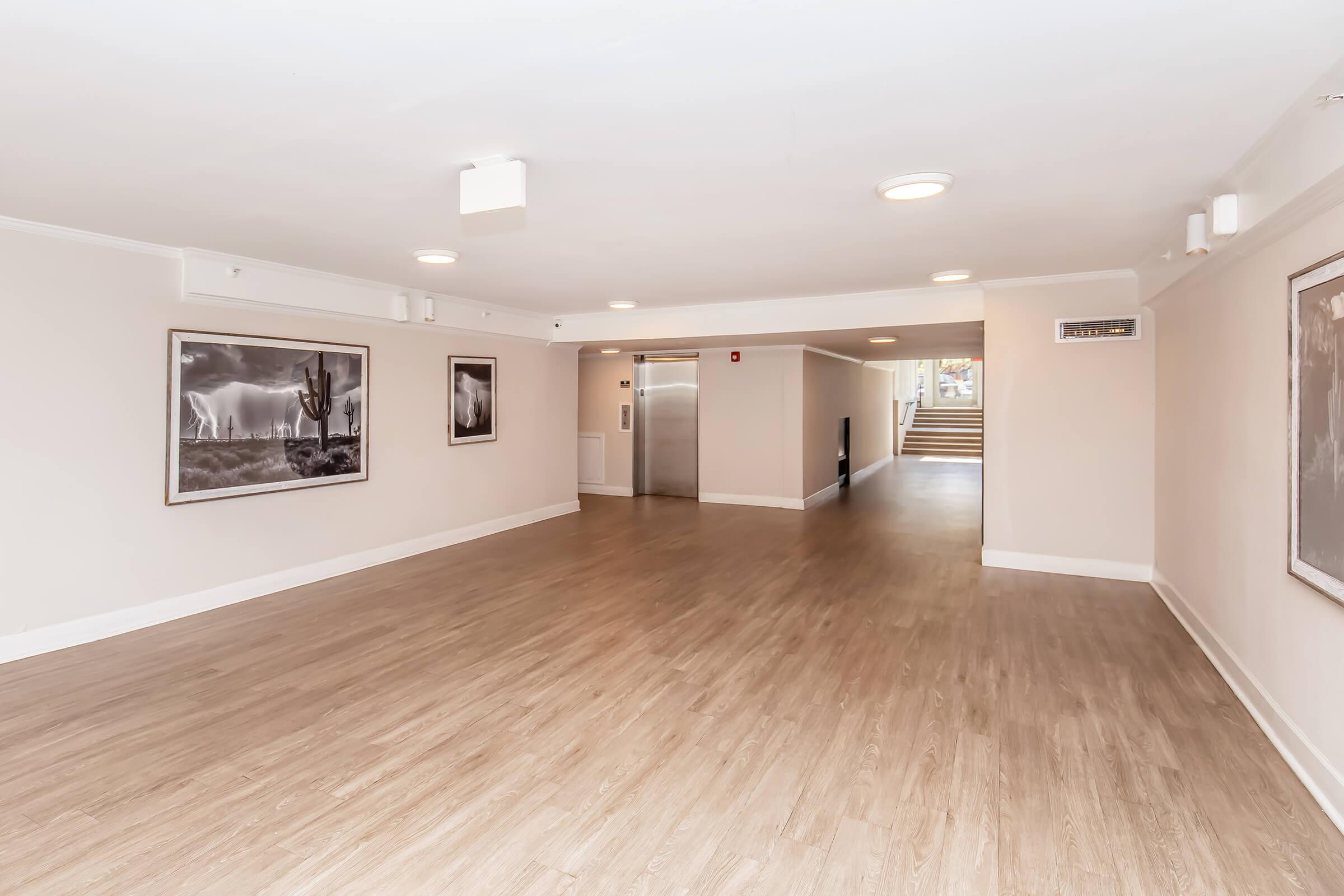 A large, empty indoor hallway featuring wooden floors, soft lighting, and beige walls. On the left, two framed monochrome photographs are displayed. An elevator is visible on the far wall, and stairs lead to another area in the background. The space appears well-maintained and inviting.