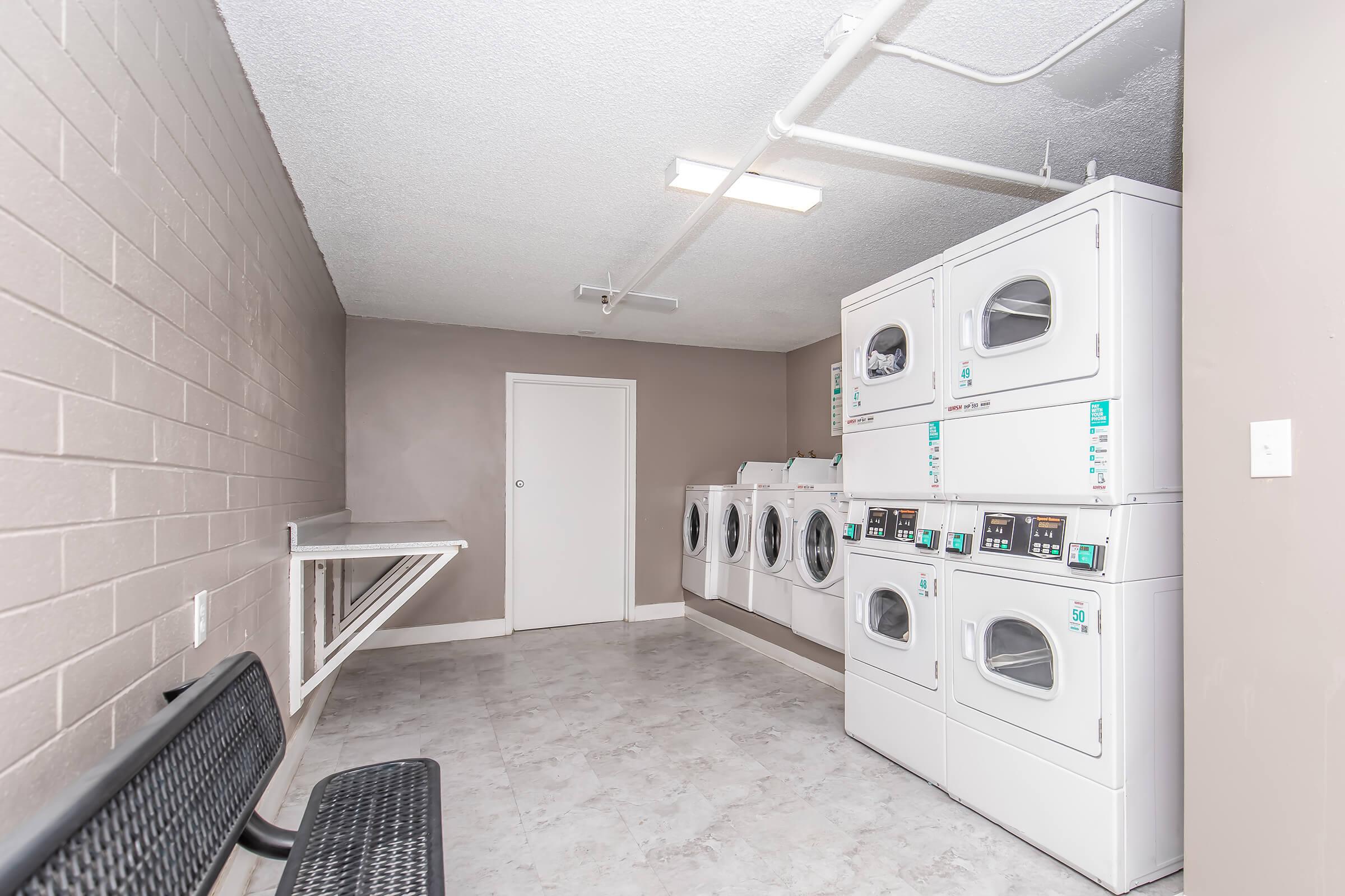 A clean, well-lit laundry room featuring multiple white washing machines and dryers arranged against a wall. There's a folding table on the side and a black bench, with gray walls and a tiled floor, creating a tidy and functional space for laundry activities.