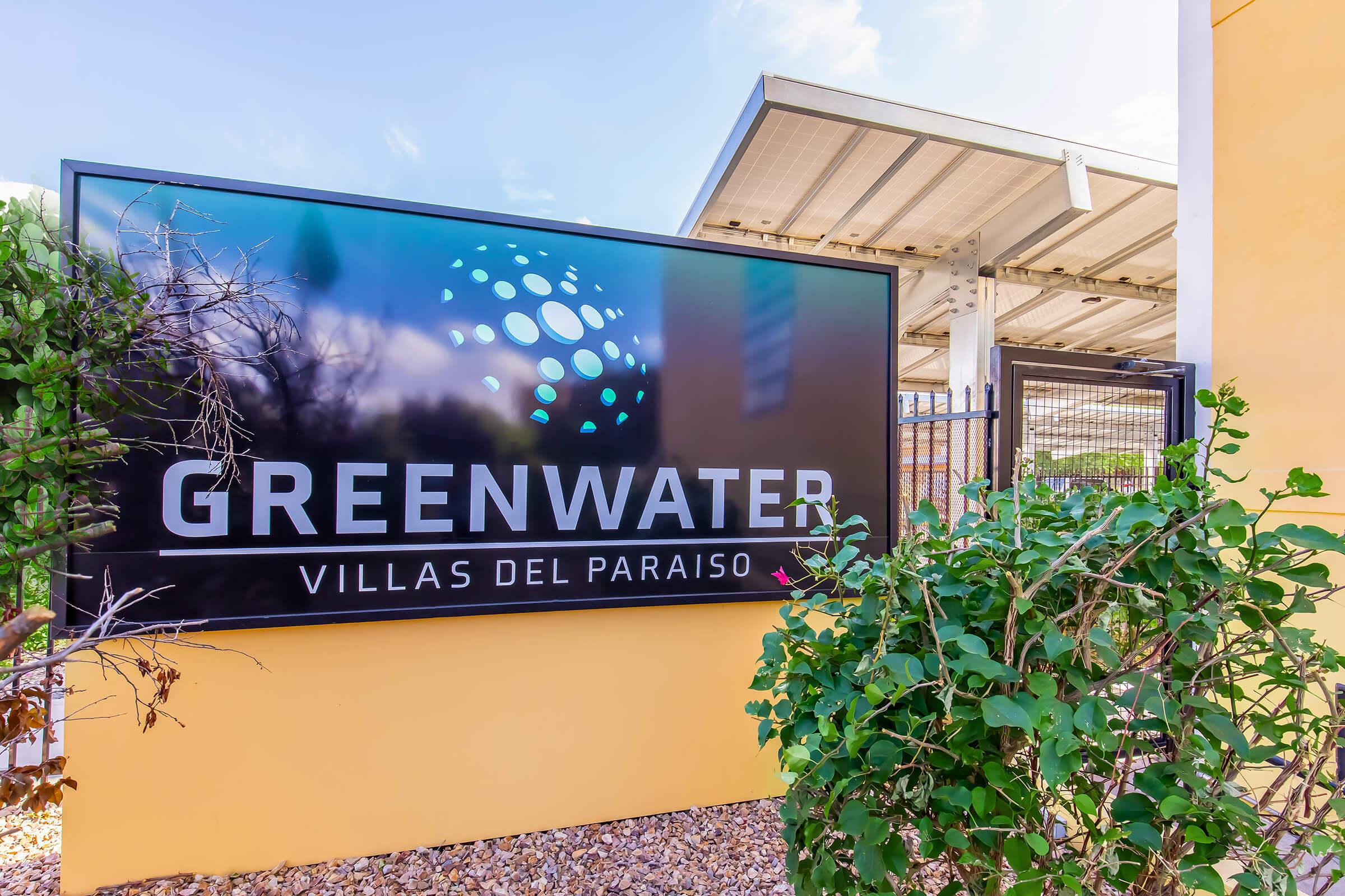Sign displaying "Greenwater Villas del Paraíso" with a modern logo, surrounded by greenery and a fence. The background features a solar panel structure and a bright sky.