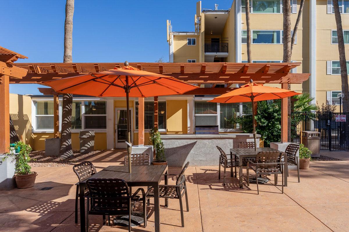 Outdoor seating area featuring several tables and chairs beneath bright orange umbrellas. In the background, a multi-story building with a yellow exterior is visible, alongside palm trees and decorative plants, under a clear blue sky. Ideal for relaxation or dining outside.