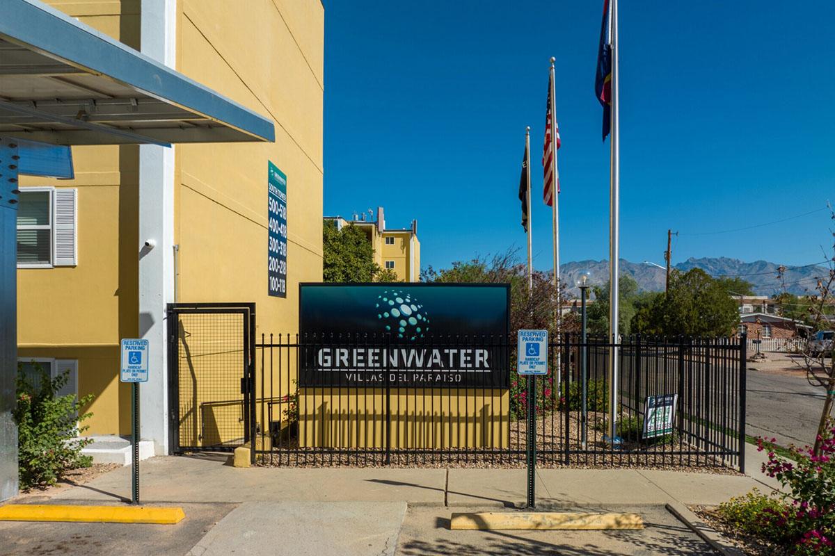 Sign for Greenwater Villas del Pafatio, featuring a logo with abstract design, surrounded by a gated entrance. Flags and mountains are visible in the background, with a clear blue sky. The building has a yellow exterior and pavement leading to the entrance.