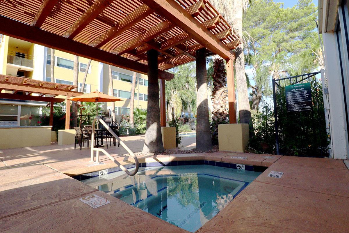 A cozy outdoor space featuring a small hot tub surrounded by wooden pergolas, tropical plants, and seating areas. Sunlight filters through, creating a warm atmosphere. In the background, a building with large windows and a pool area can be seen.