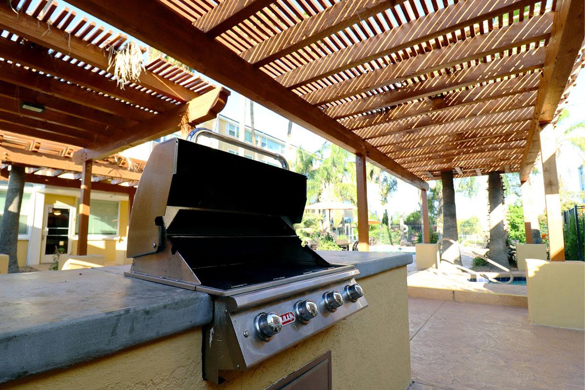 A stainless steel gas grill situated in an outdoor space with a wooden pergola overhead. The area features palm trees and a paved surface, creating a comfortable setting for barbecues and gatherings.
