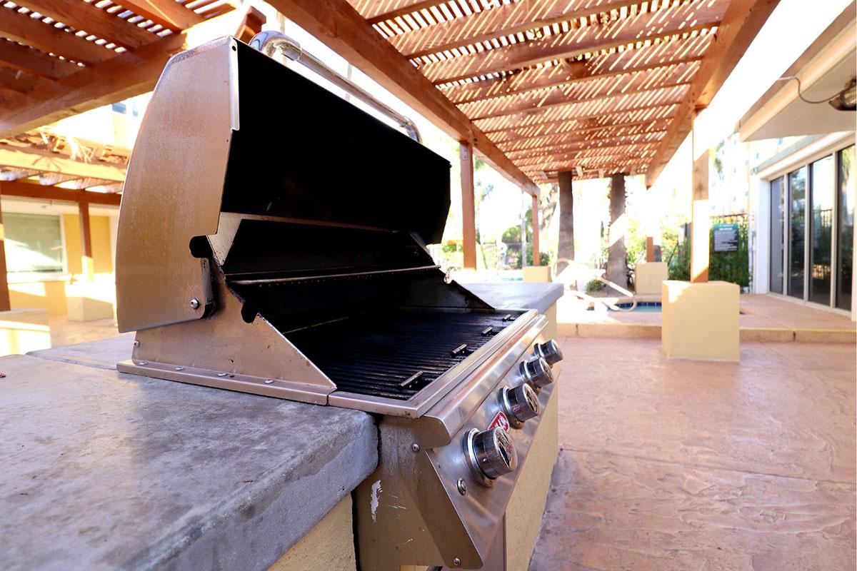 A stainless steel outdoor grill with an open lid, showcasing a clean cooking surface. The grill is situated under a wooden pergola, with a sunny patio and palm trees in the background, indicating a relaxing outdoor space ideal for gatherings and barbecues.