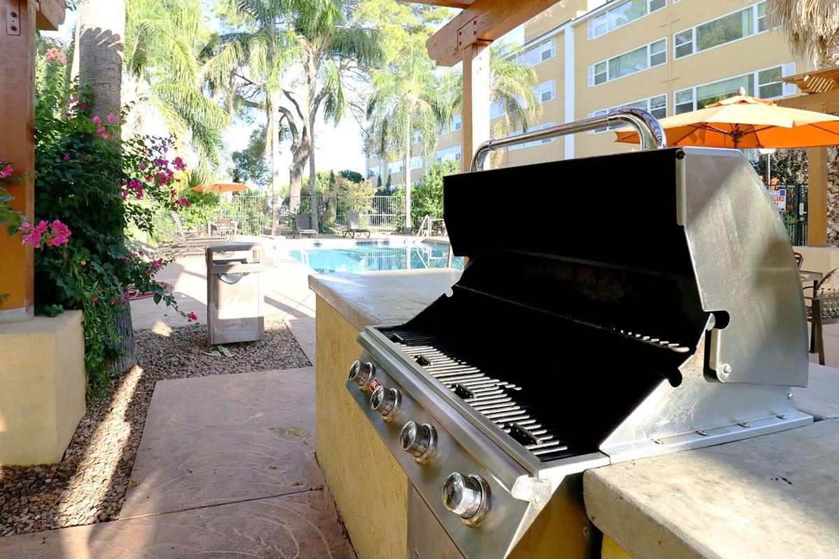 A stainless steel gas grill positioned near a swimming pool, surrounded by tropical palm trees and colorful flowers. Patio umbrellas are visible in the background, creating a lively outdoor atmosphere. The grill appears clean and ready for use.