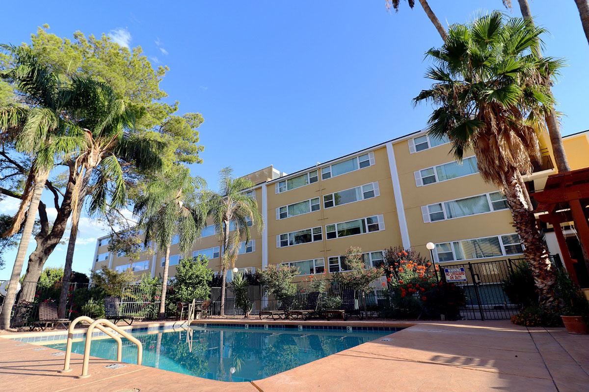 A colorful, multi-story building with several windows is located near a swimming pool, surrounded by palm trees and landscaped greenery. The sky is clear and blue, creating a sunny and inviting atmosphere. The pool area features a few lounge chairs and a safety fence.