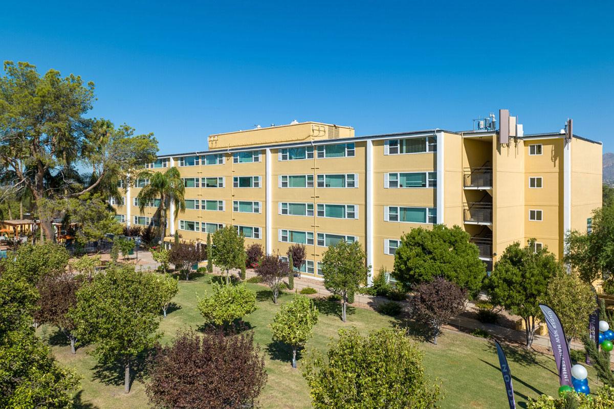 A modern yellow multi-story building surrounded by green trees and well-maintained landscaping. The structure features balconies and large windows, reflecting a clear blue sky. Nearby, there are patches of various types of vegetation, indicating a serene outdoor environment.