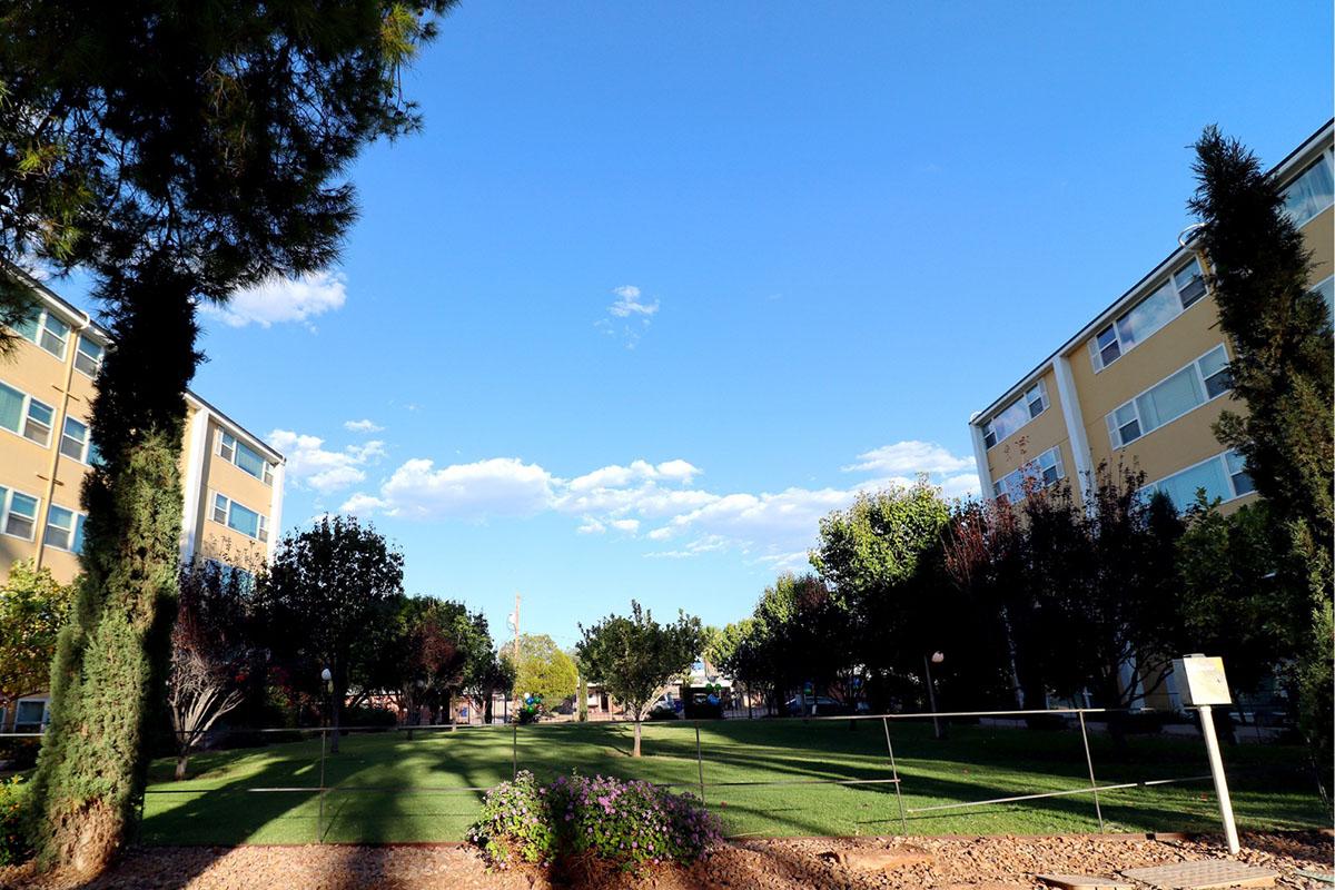A view of a landscaped area between two apartment buildings, featuring green grass, trees, and bushes under a blue sky with scattered clouds. The scene conveys a peaceful residential environment.
