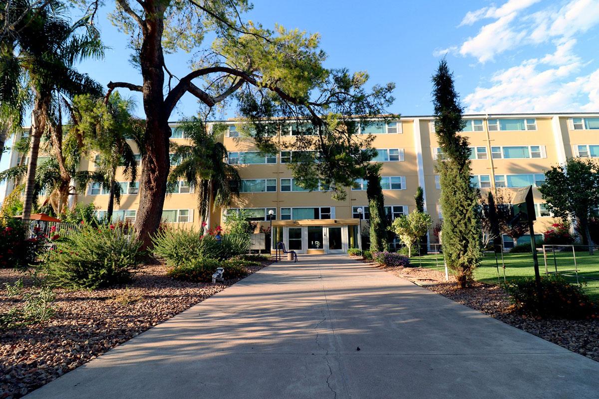 A pathway leads to a modern, multi-story building surrounded by various greenery, including palm trees and shrubs. The sky is partly cloudy, and the setting appears to be well maintained with an inviting atmosphere.