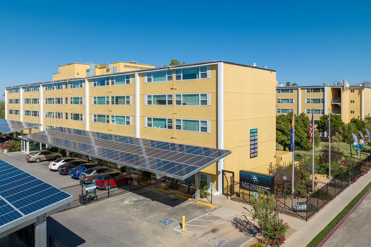A modern multi-story building with a yellow exterior featuring solar panels on the roof. The foreground shows a parking area with several cars, and there are green landscaped areas with trees and plants. The sky is clear and blue, contributing to an eco-friendly atmosphere.