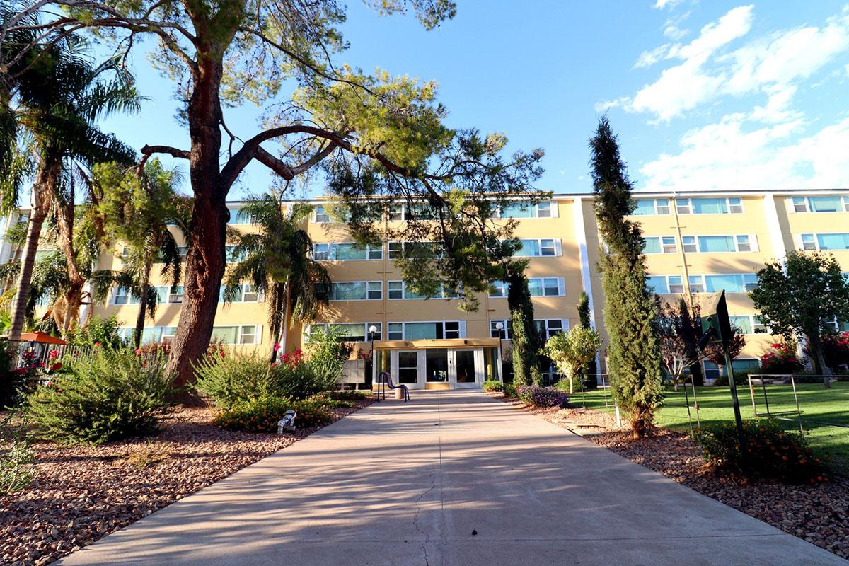 A pathway leading to a yellow building surrounded by palm trees and greenery. The building features multiple windows and is two stories high, with a clear blue sky in the background. The area is well landscaped with shrubs and plants, creating a welcoming entrance.