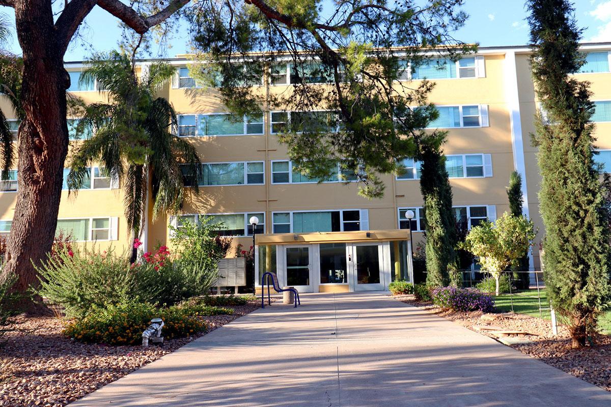 A modern three-story building surrounded by trees and colorful landscaping. A wide path leads to the entrance, which features glass doors and outdoor seating. Lush plants and flowering shrubs adorn the area, creating a welcoming atmosphere.