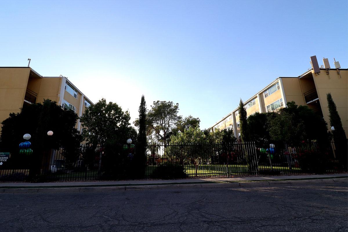 A view of two apartment buildings facing each other, separated by a green landscaped area with trees. The foreground features a gated entrance and decorative lights, while the background showcases a clear blue sky. Shadows of the buildings fall across the pavement, creating a serene urban atmosphere.