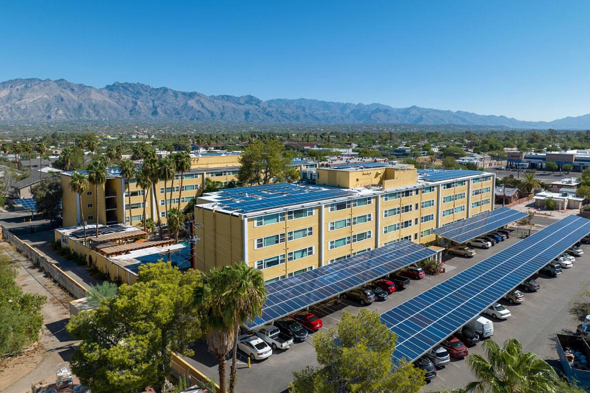 Aerial view of a large yellow building with solar panels on the roof, surrounded by palm trees. In the foreground, a parking area with several cars is shaded by solar panel canopies. Desert mountains are visible in the background against a clear blue sky.