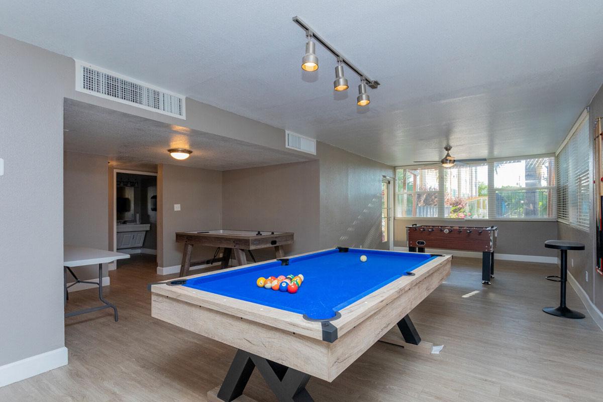 A recreation room featuring a blue pool table with colorful balls arranged on it, a foosball table, and seating. The walls are painted in neutral colors, and there's good lighting from overhead fixtures and natural light coming through windows.
