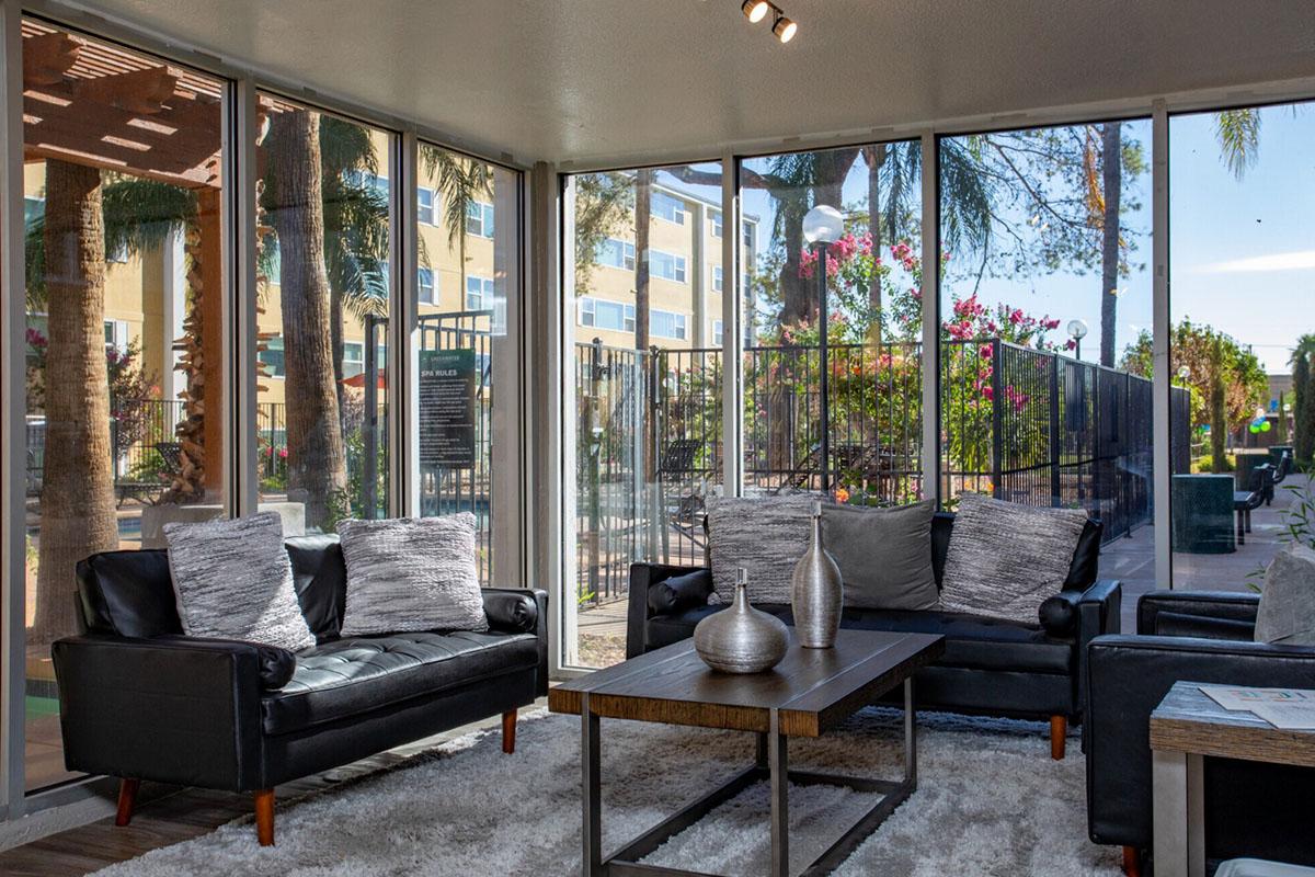 A bright, modern living space featuring two black couches with decorative pillows, a wooden coffee table, and a soft rug. The room has large windows showcasing a landscaped outdoor area with palm trees and flowers, allowing natural light to fill the space.