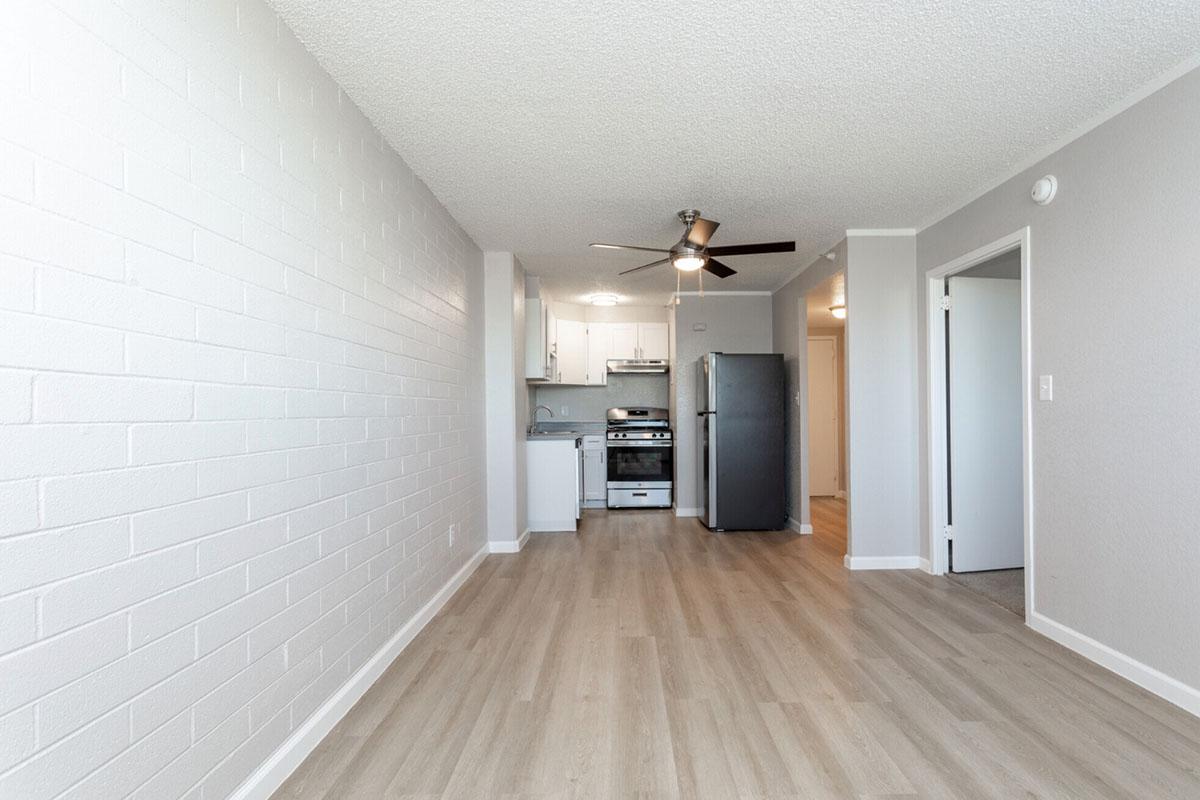 Interior view of a modern, open-concept apartment featuring light-colored walls, a spacious living area with laminate flooring, and a kitchen with white cabinets, stainless steel appliances, and a ceiling fan. The layout allows for natural light, giving a bright and airy feel to the space.