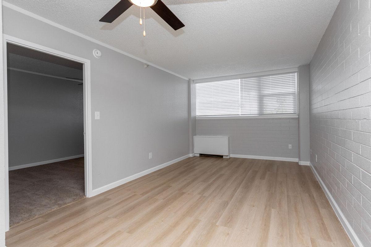 A sparsely furnished interior room with light gray walls and ceiling. There is a ceiling fan, a window with blinds allowing natural light, and a heater on the wall. The flooring is light-colored laminate, and an open doorway leads to another room with carpet. Overall, the space appears clean and modern.