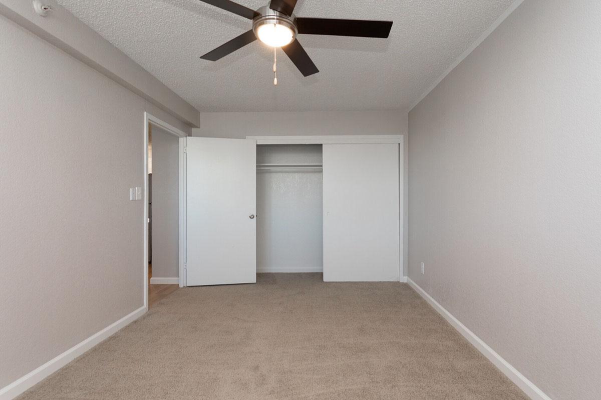 An empty bedroom featuring light gray walls, a ceiling fan, and beige carpet. There are two closed white doors leading to a closet on the left side, and a doorway on the right that possibly leads to another room. The lighting is bright, creating an inviting atmosphere.