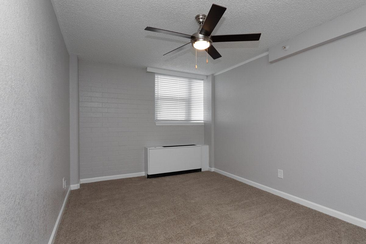 A vacant room featuring light gray walls, a ceiling fan with five blades, and a window with blinds. The floor is covered in beige carpet, and there is a baseboard heater along one wall. The space is well-lit and has a minimalistic design, suitable for various furniture arrangements.