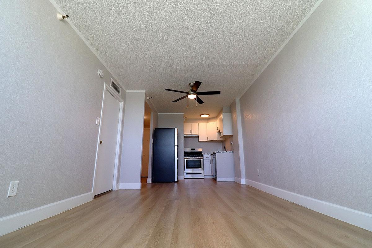View of a modern apartment interior featuring a spacious living area with light-colored wooden flooring. A kitchen area is visible in the background, equipped with a black refrigerator, white cabinets, and a stove. The ceiling has a modern fan, and walls are painted in neutral tones, creating an open and airy atmosphere.