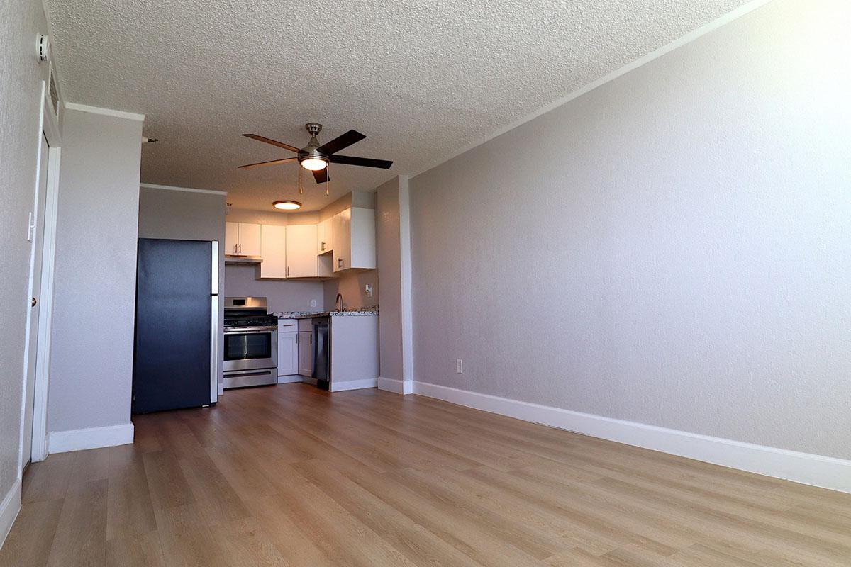 Spacious apartment interior featuring light-colored walls and wooden flooring. A modern kitchen area with white cabinetry and stainless steel appliances is visible, along with a ceiling fan. The room has ample natural light, creating an inviting atmosphere.
