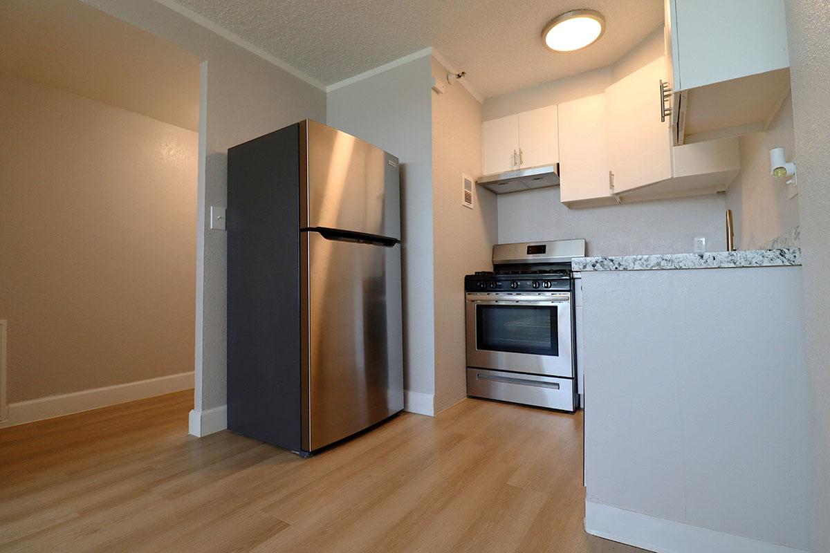 A modern kitchen featuring stainless steel appliances, including a refrigerator and gas stove. White cabinets and a granite countertop complement the light wood flooring, creating a bright and inviting space. The image captures the kitchen from a corner angle, showing an open layout.