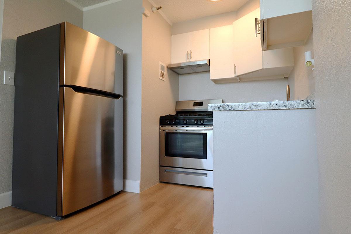 A modern kitchen featuring a stainless steel refrigerator, a gas stove, and an oven, with white cabinetry and a granite countertop. The floors are light-colored, and the walls are painted in a neutral tone, creating a bright and clean atmosphere.