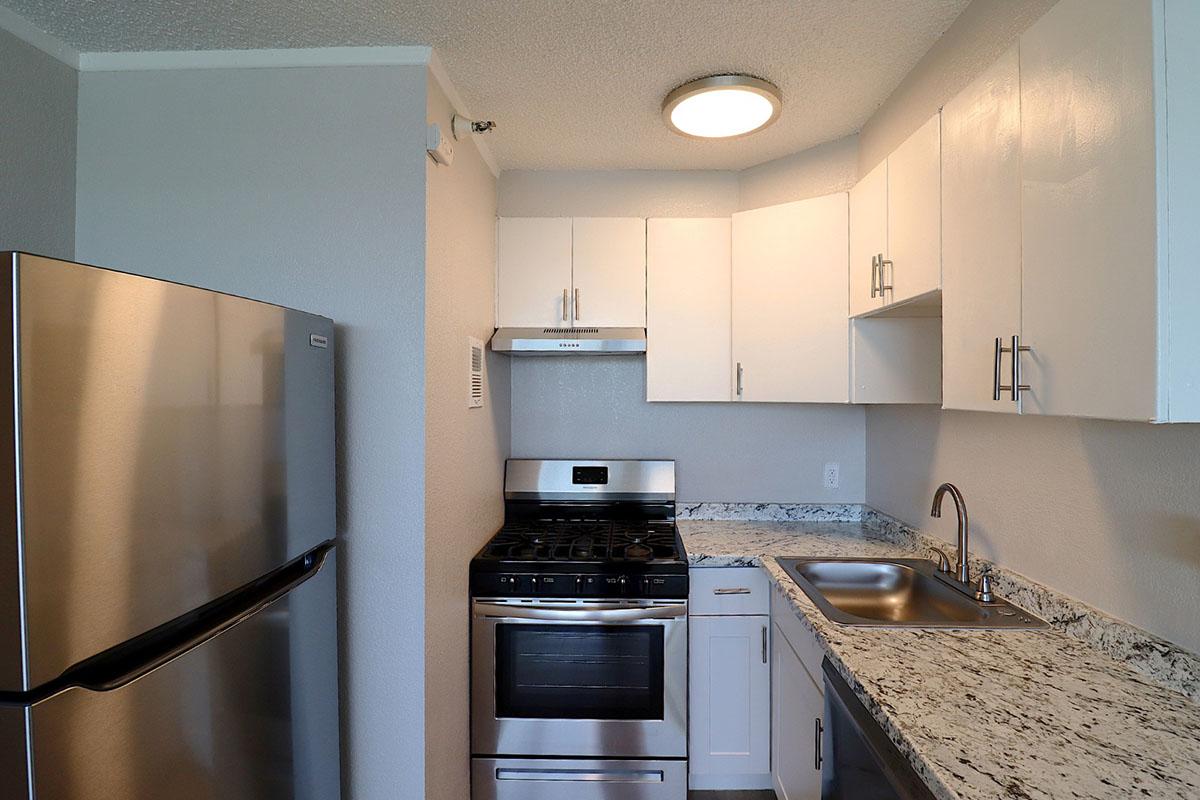Modern kitchen featuring stainless steel appliances, including a refrigerator, gas stove, and oven. The countertops are made of granite, and there are white cabinets above and below. The walls are painted in a light gray color, and the area is well-lit with a ceiling light.