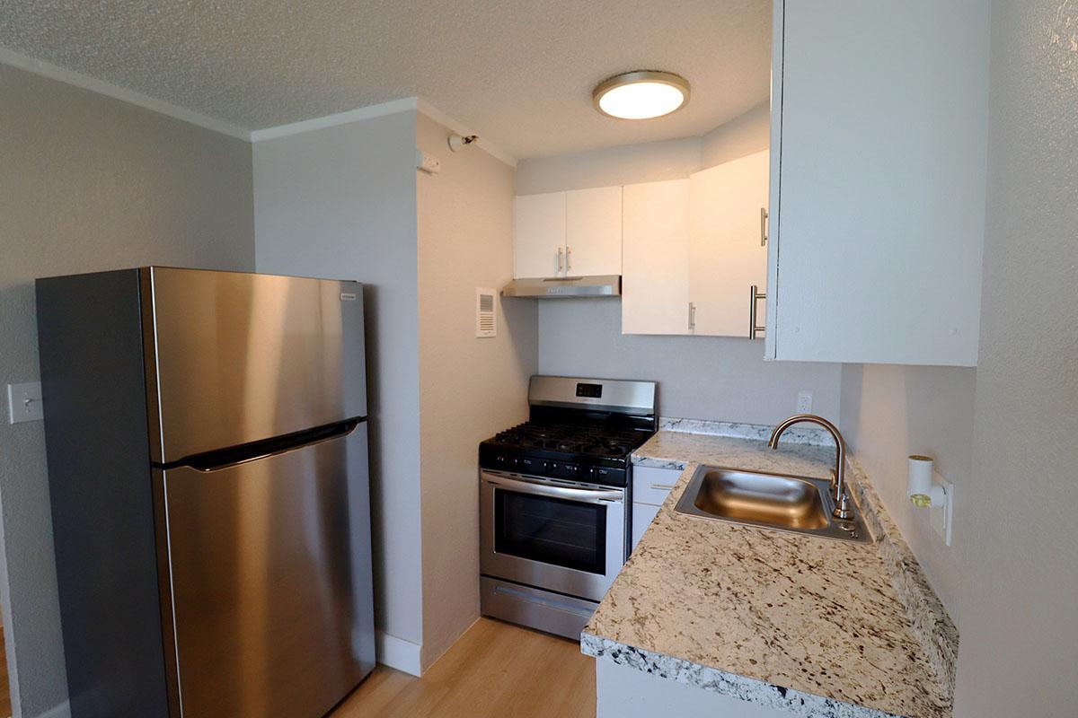A modern kitchen featuring a stainless steel refrigerator, a gas stove with an oven, and a sink. The cabinetry is white, and there’s a granite countertop. The walls are light gray, and a ceiling light fixture provides illumination. The layout is compact and functional.