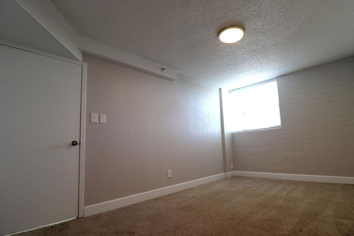 A spacious and well-lit room featuring beige carpet, a textured ceiling with a circular light fixture, white walls, and a window allowing natural light. A closed white door is visible on the left, while the right side shows a bare wall. The overall atmosphere is neutral and inviting.