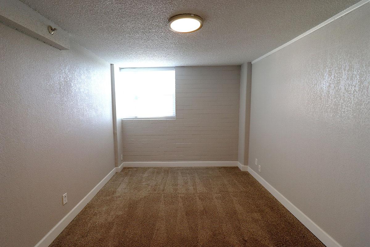 A small, empty room with light beige carpet and light gray walls. There is a window on one side, allowing natural light to enter, and a simple ceiling light fixture. The room appears clean and ready for furniture placement.