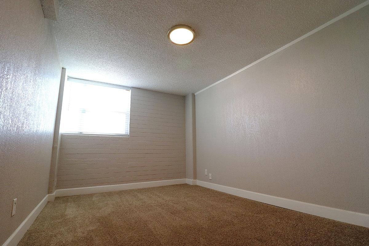 A small, empty room with a light gray textured wall and light brown carpet. There is a window allowing natural light to enter, and a ceiling light fixture. The overall space appears clean and minimalistic, suitable for various uses such as a bedroom or office.