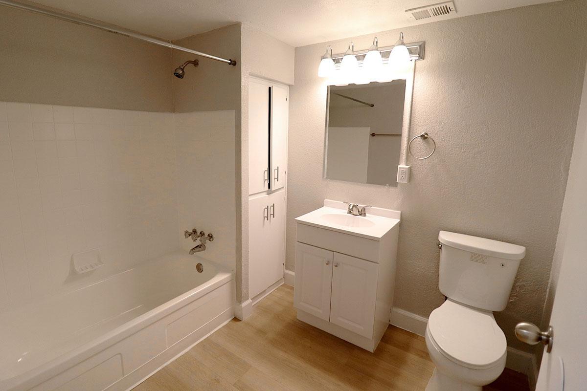 A modern bathroom featuring a white bathtub with a shower, a compact vanity with a sink and mirror, a toilet, and light-colored walls. The floor is designed with light-colored wood laminate, and there is a towel ring on the wall next to the mirror.