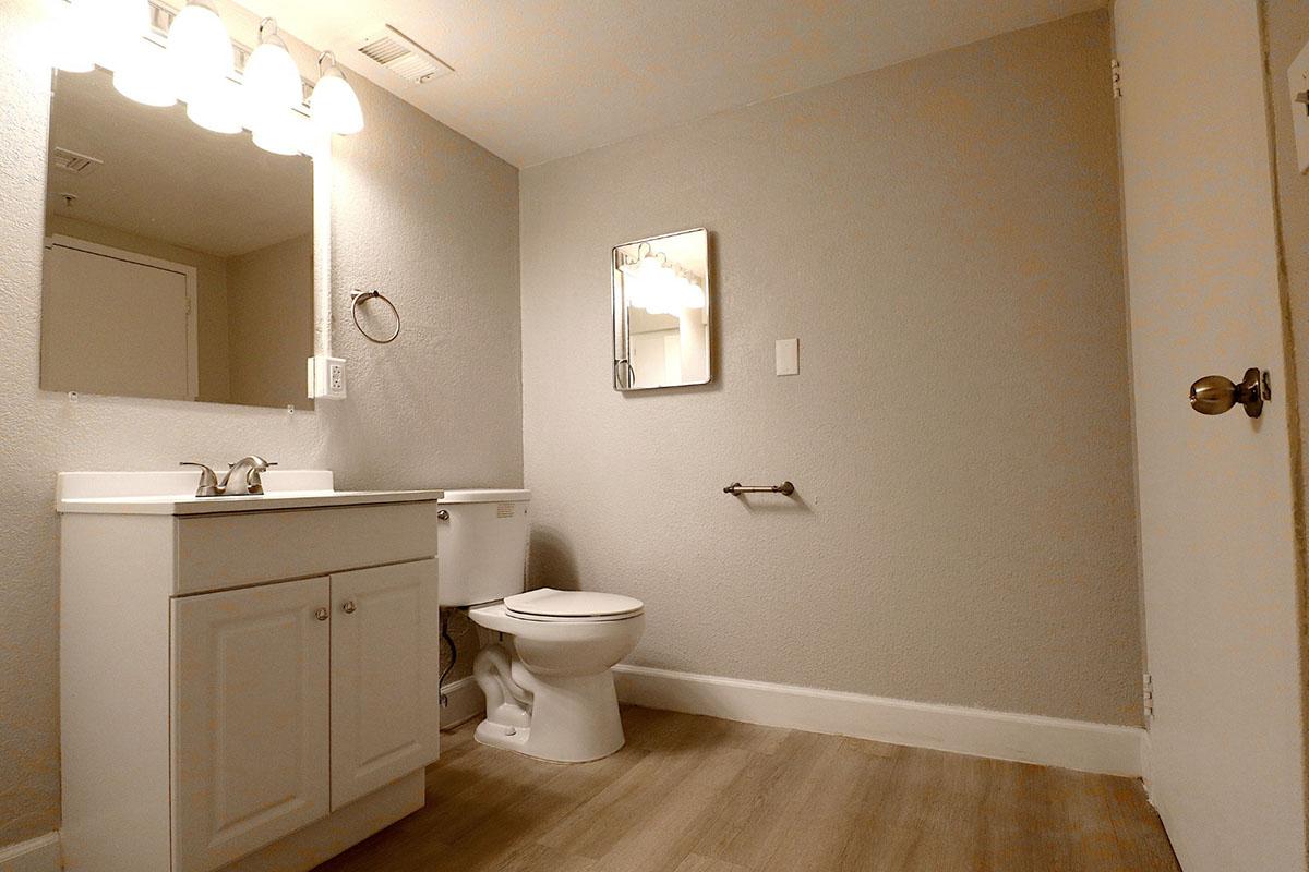 A small, modern bathroom featuring a white vanity with a sink, a mirror above it, a toilet, and a towel rack. The walls are painted light gray, and the floor has a light wood-like design. Three light fixtures illuminate the space from above.