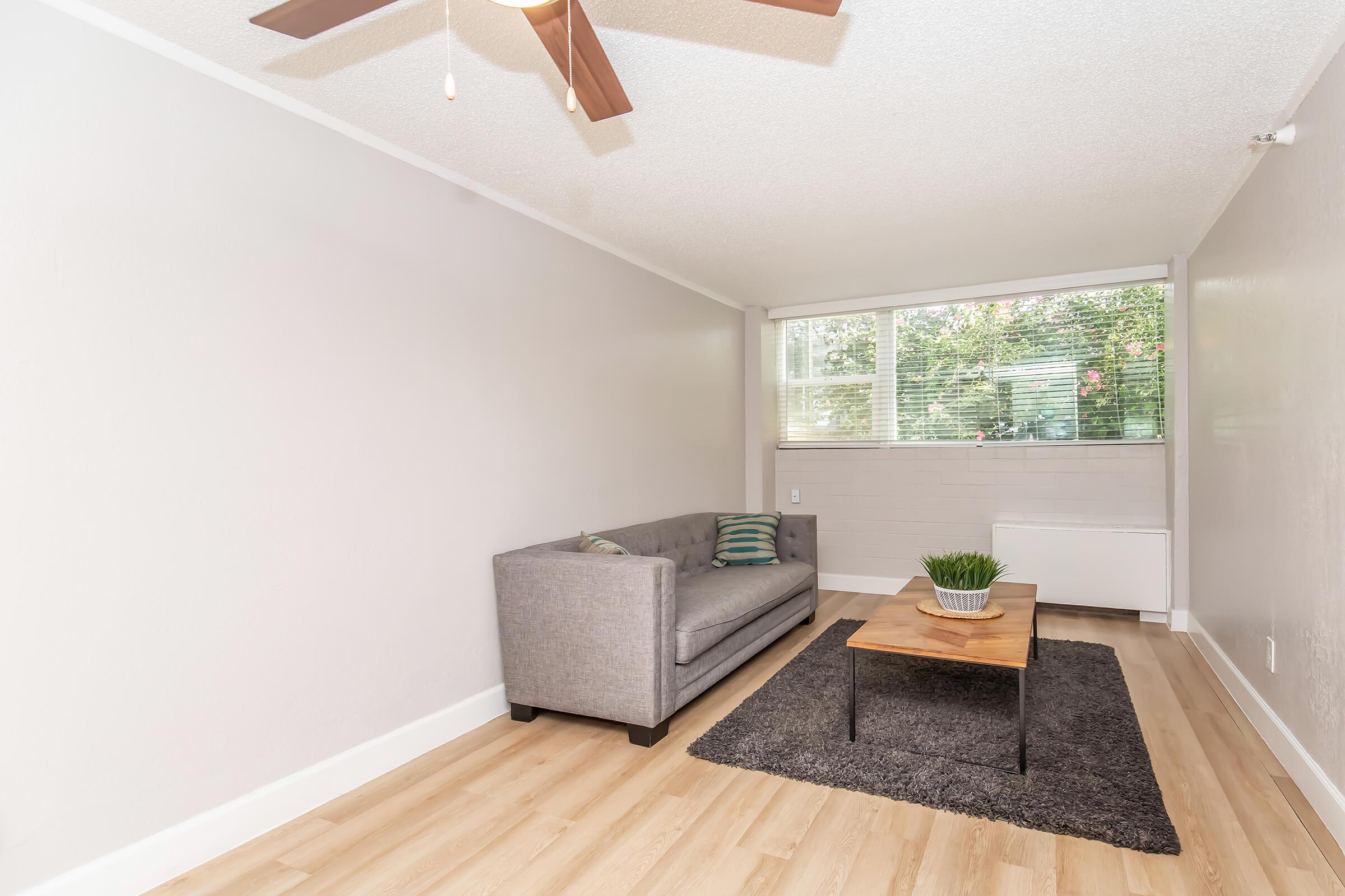 A cozy living room featuring a gray sofa with decorative pillows, a wooden coffee table on a dark rug, and a ceiling fan. Large windows let in natural light and greenery from outside is visible. The walls are light-colored and the flooring is wooden.