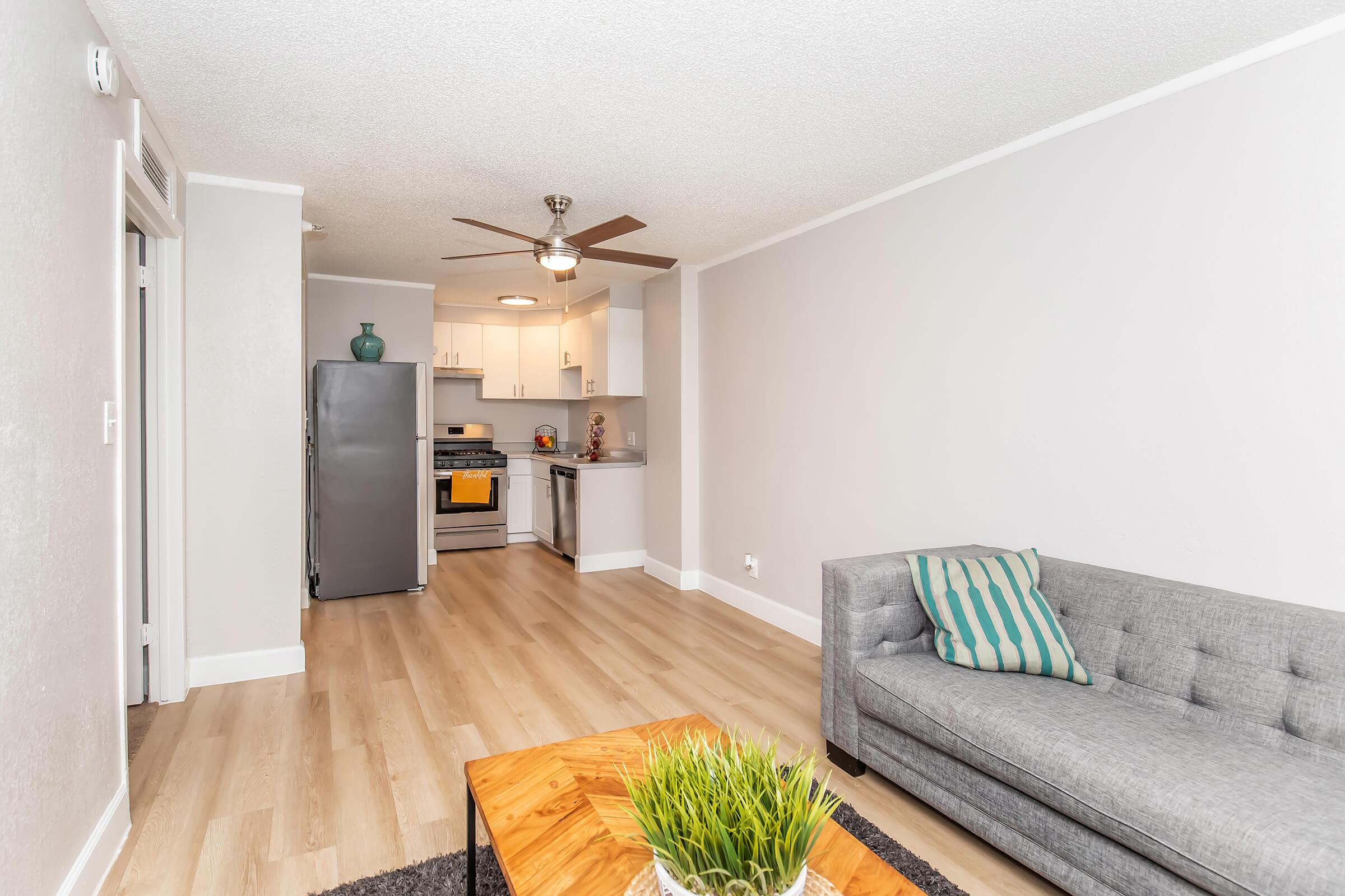 Interior of a modern apartment featuring a light gray living room with a comfortable gray sofa and an accent table with a plant. The kitchen is visible in the background, showcasing white cabinets and stainless steel appliances, including a refrigerator. Warm wood flooring complements the space.