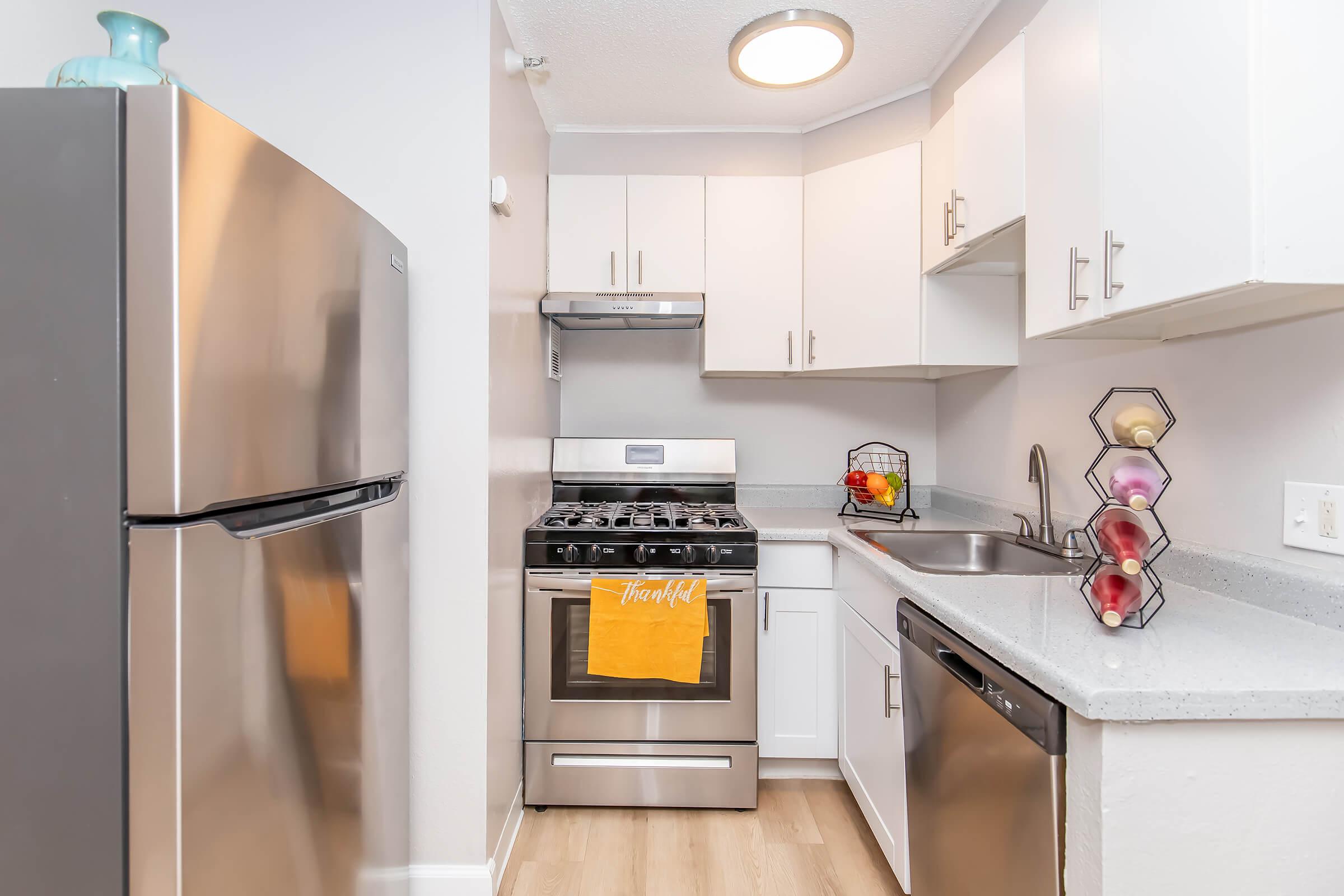 A modern kitchen featuring stainless steel appliances, including a refrigerator and gas stove. The cabinets are white, and there's a gray countertop. A sink is situated next to the stove, with a dish rack holding colorful fruits. A yellow dish towel hangs on the stove. The floor has light wood-like laminate.