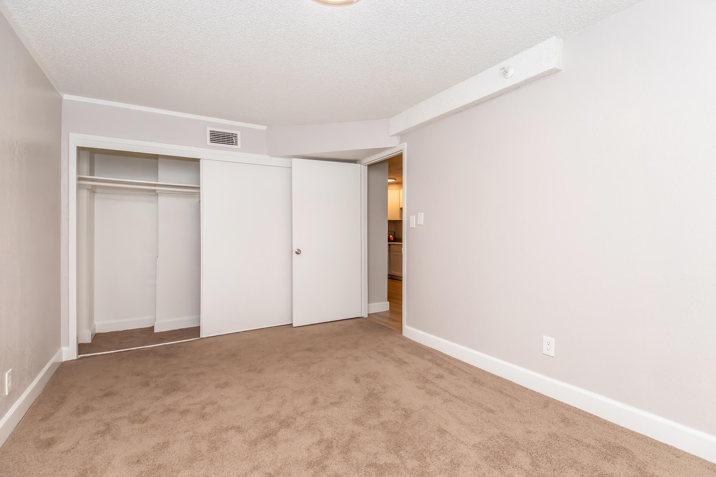 A spacious empty room with light beige carpet and light gray walls. There is a closet with sliding doors on the left side and an open doorway leading to another area at the back. Natural light is coming from the entrance, enhancing the airy feel of the space.
