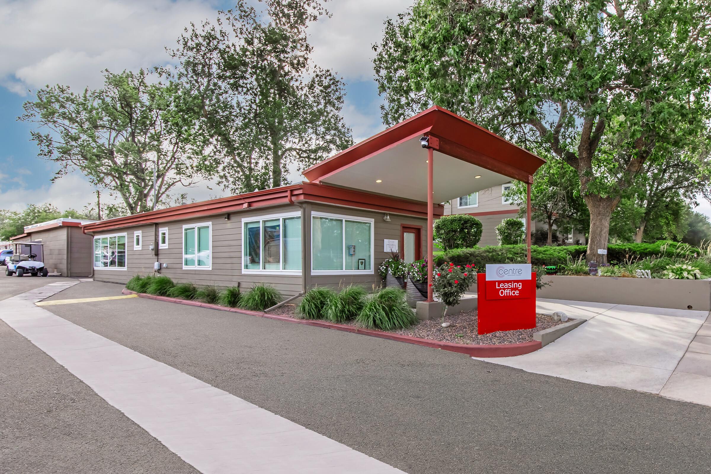 A leasing office building with a modern design, featuring large windows and a red roof. It has a prominent red sign reading "Leasing Office" in front. Surrounding the building are lush green plants and trees, with a paved path leading up to the entrance. The area appears well-maintained and inviting.