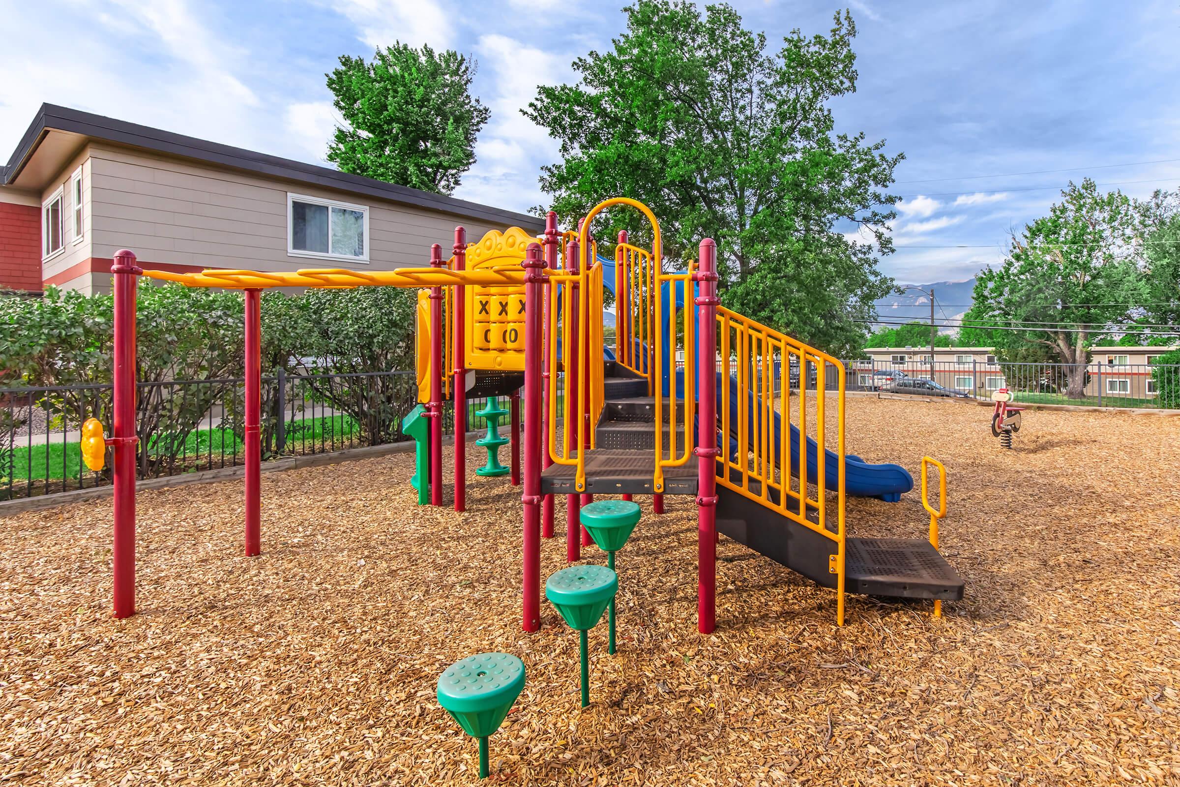 A colorful playground featuring a slide, climbing structure, and swing set, surrounded by wood mulch and green trees. The area is sunny with a clear sky, and a nearby house is visible in the background.