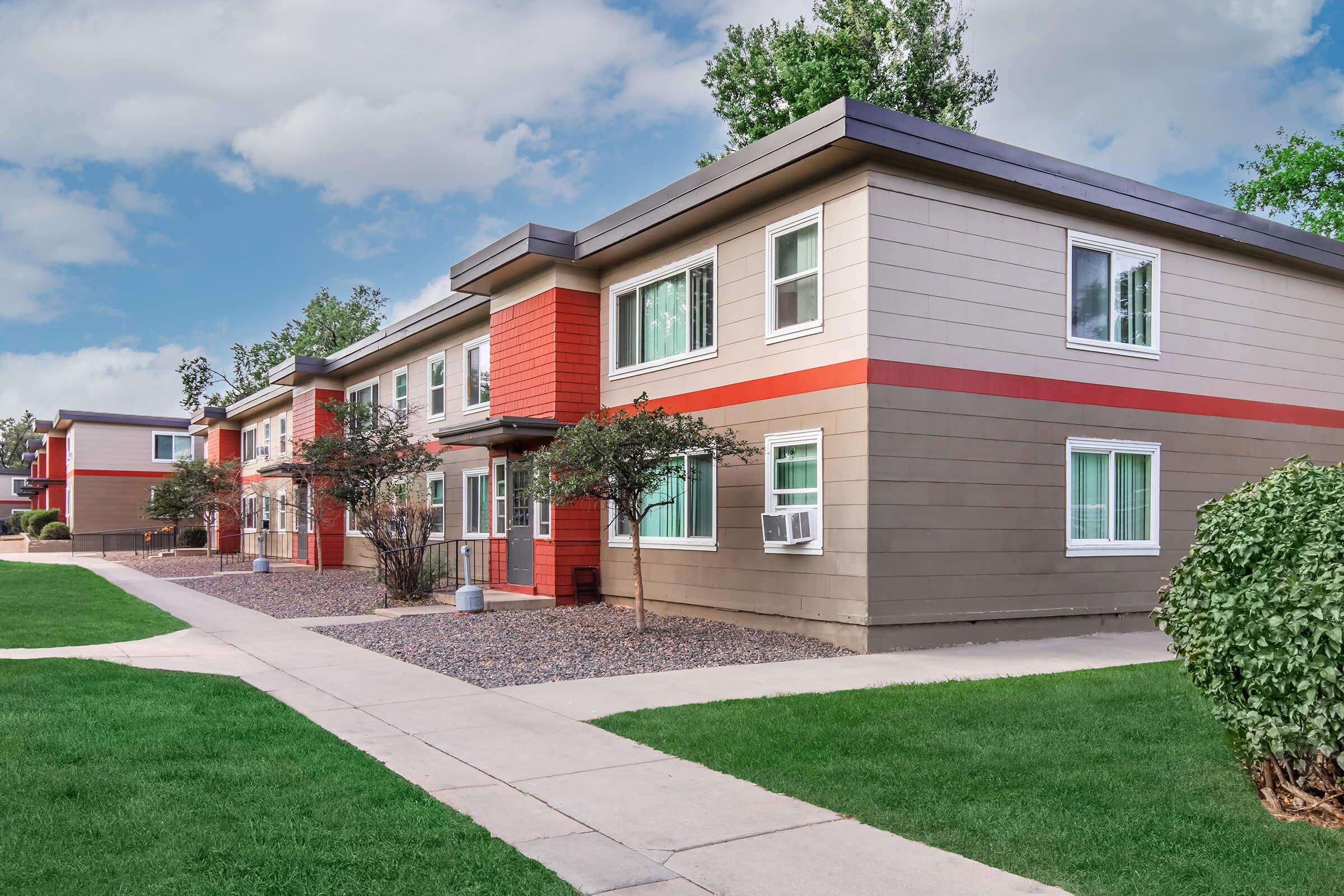 Two-story apartment building with a symmetrical design featuring a mix of red and gray siding. The exterior includes several windows, and there are small trees and shrubs in front of the building. A walkway leads to the entrance, with a grassy lawn in the foreground and a cloudy sky above.