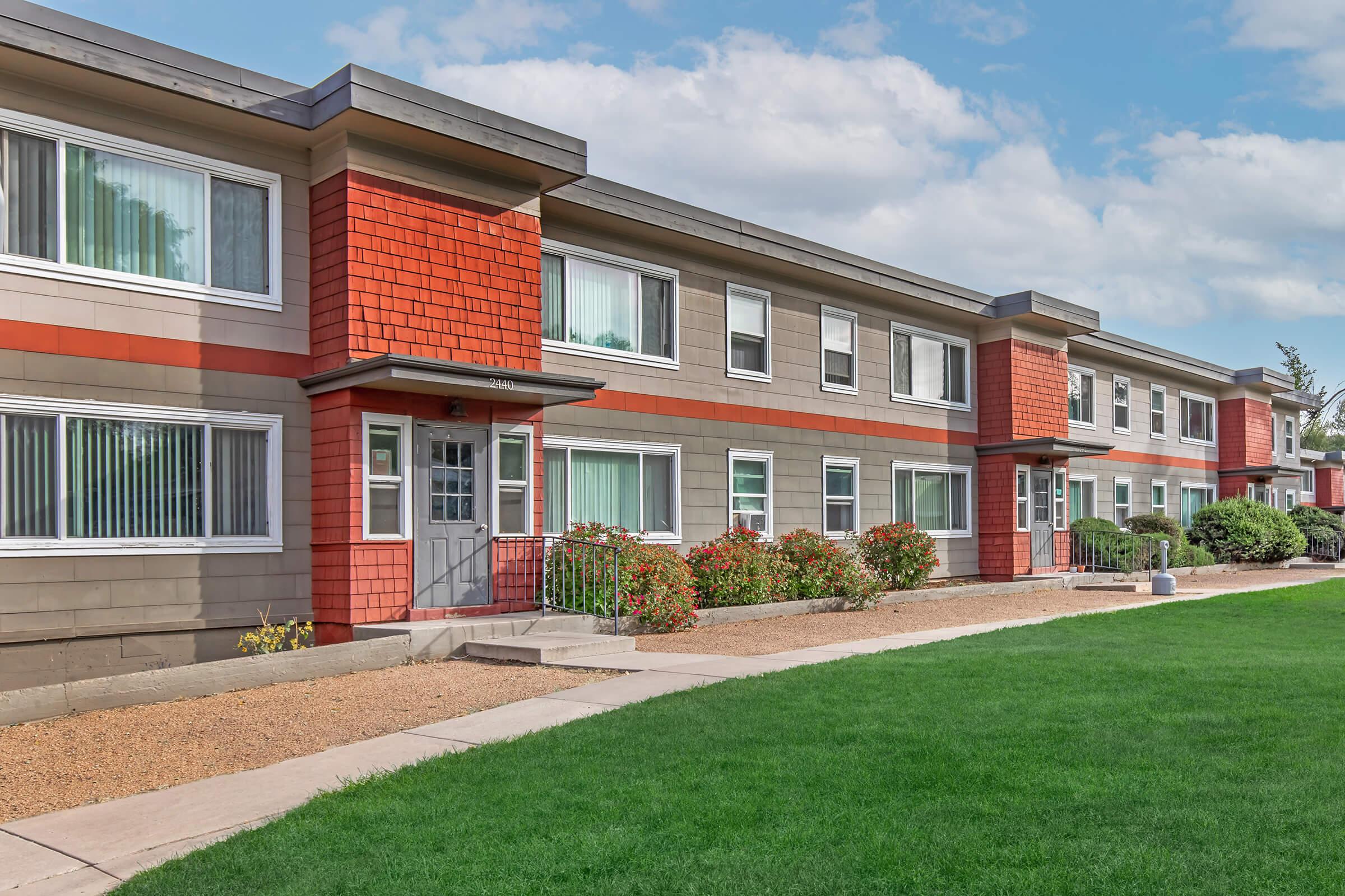 Two-story apartment building featuring a mix of brown and red siding. The facade includes multiple windows and a central door, with well-maintained landscaping including a green lawn, flowers, and shrubs in front. The sky is partly cloudy, adding to the overall pleasant ambiance of the scene.
