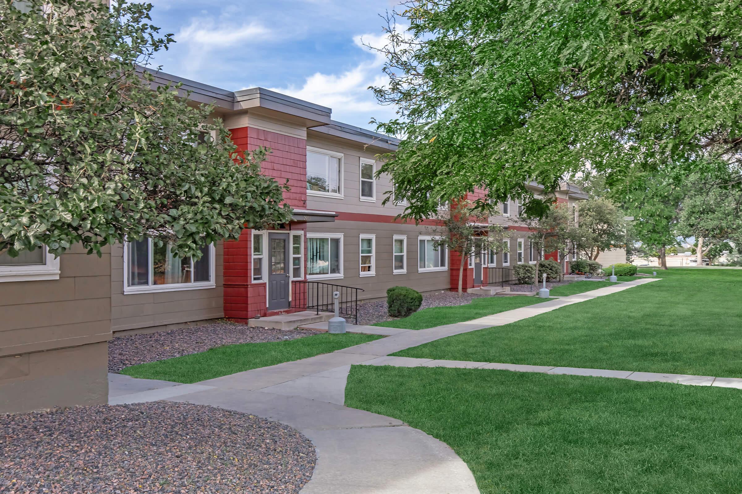 A modern apartment complex featuring multiple buildings with a combination of beige and red facades. The landscape includes green lawns, neatly trimmed shrubs, and a paved walkway leading to the entrances. Trees provide shade, and the sky appears bright and clear in the background.