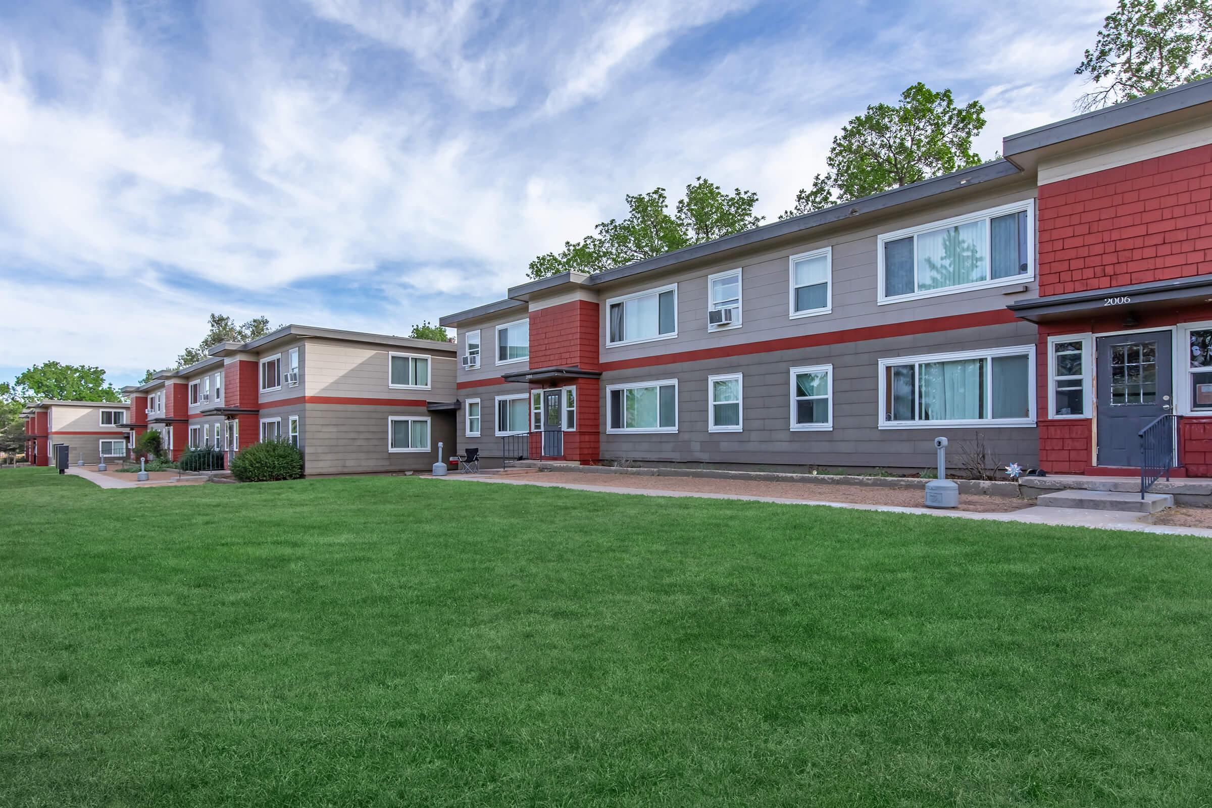 A row of modern, two-story apartment buildings with a mix of red and gray siding, set against a blue sky with scattered clouds. The well-maintained grass in the foreground adds to the inviting atmosphere, while walking paths and plants are visible along the building's exterior.