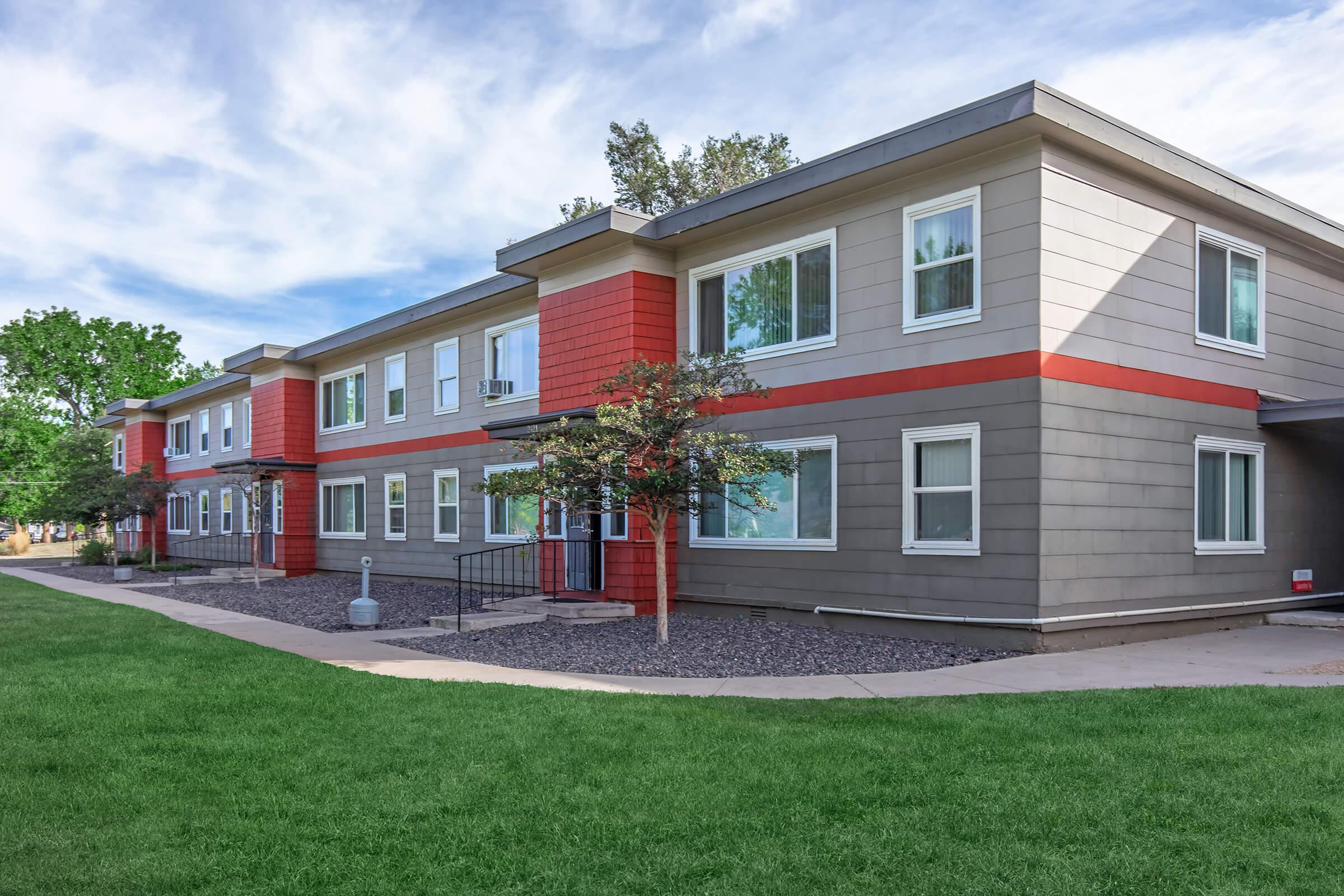 Modern multi-unit residential building with a well-maintained lawn. The building features a mix of gray and red exterior with multiple windows. A pathway runs alongside the structure, leading to the entrances. Trees and shrubs are present, contributing to a pleasant outdoor environment.